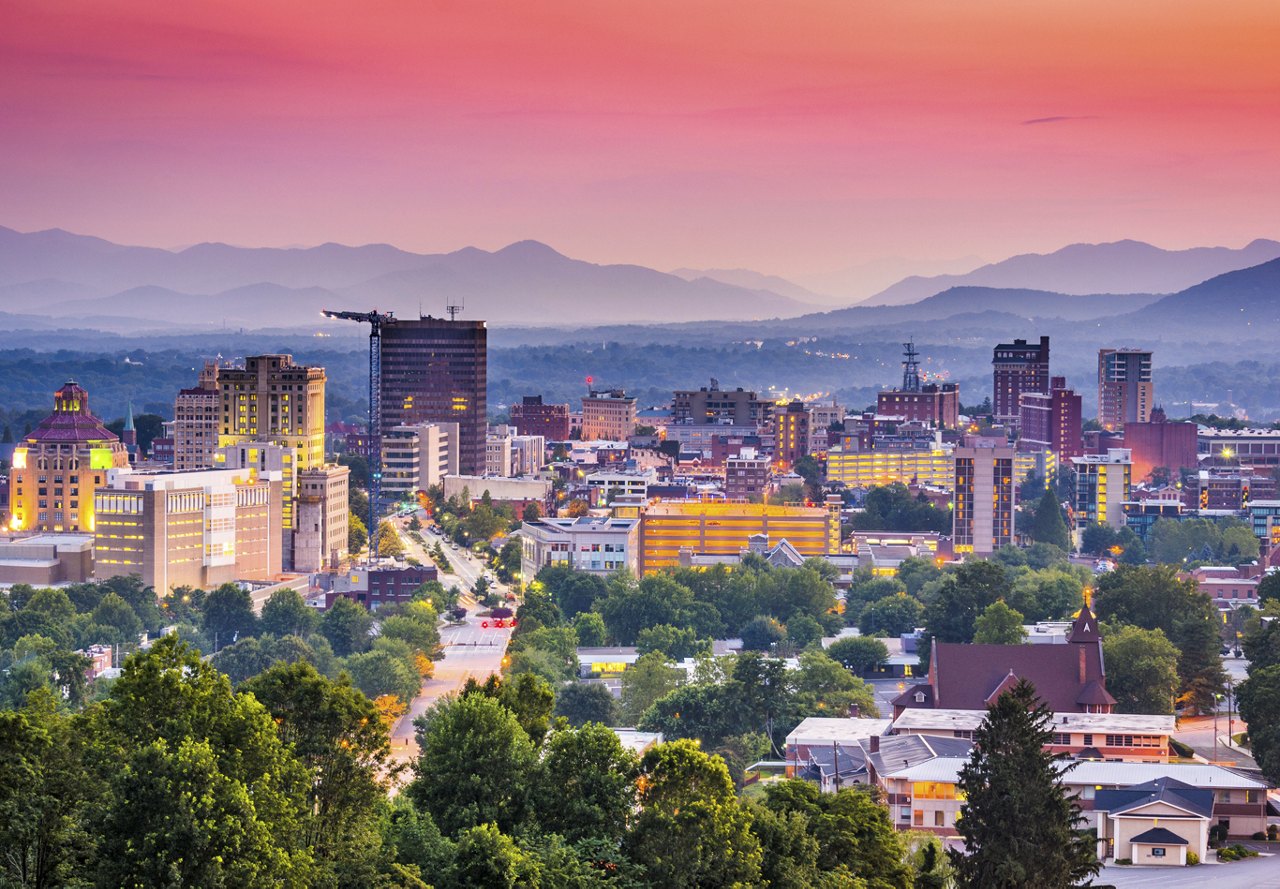 A stunning sunset in Asheville, North Carolina, with a view of the city, trees, and mountains in the background, highlighting the natural beauty of the area.