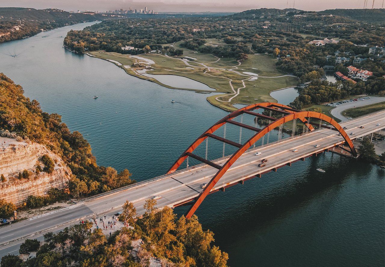 A view of a bridge in Austin, Texas, with the river flowing beneath it. The scene captures some of the surrounding buildings, and the sun creates a beautiful glow over the area.