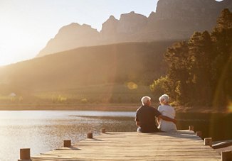 An older couple sitting at the end of a dock, enjoying each other's company and the surrounding nature. The sun shines brightly over the peaceful lake and mountains in the background, creating a serene atmosphere.