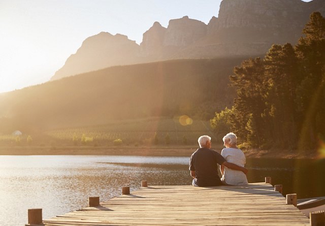 An older couple sitting at the end of a dock, enjoying each other's company and the surrounding nature. The sun shines brightly over the peaceful lake and mountains in the background, creating a serene atmosphere.