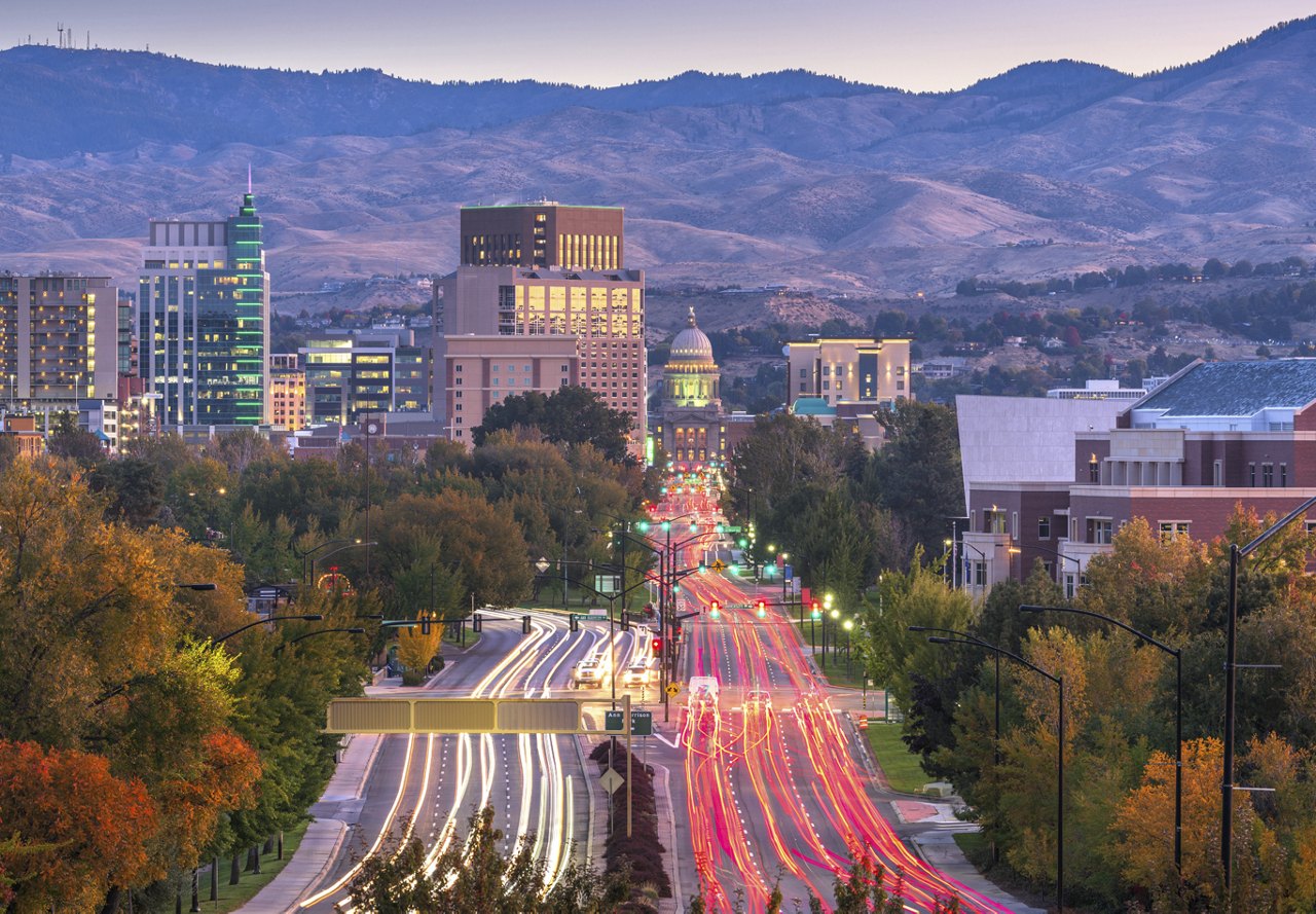 A view of major roads in Boise, Idaho, with car lights streaking through the city. The background features mountains and buildings, highlighting the vibrant area.