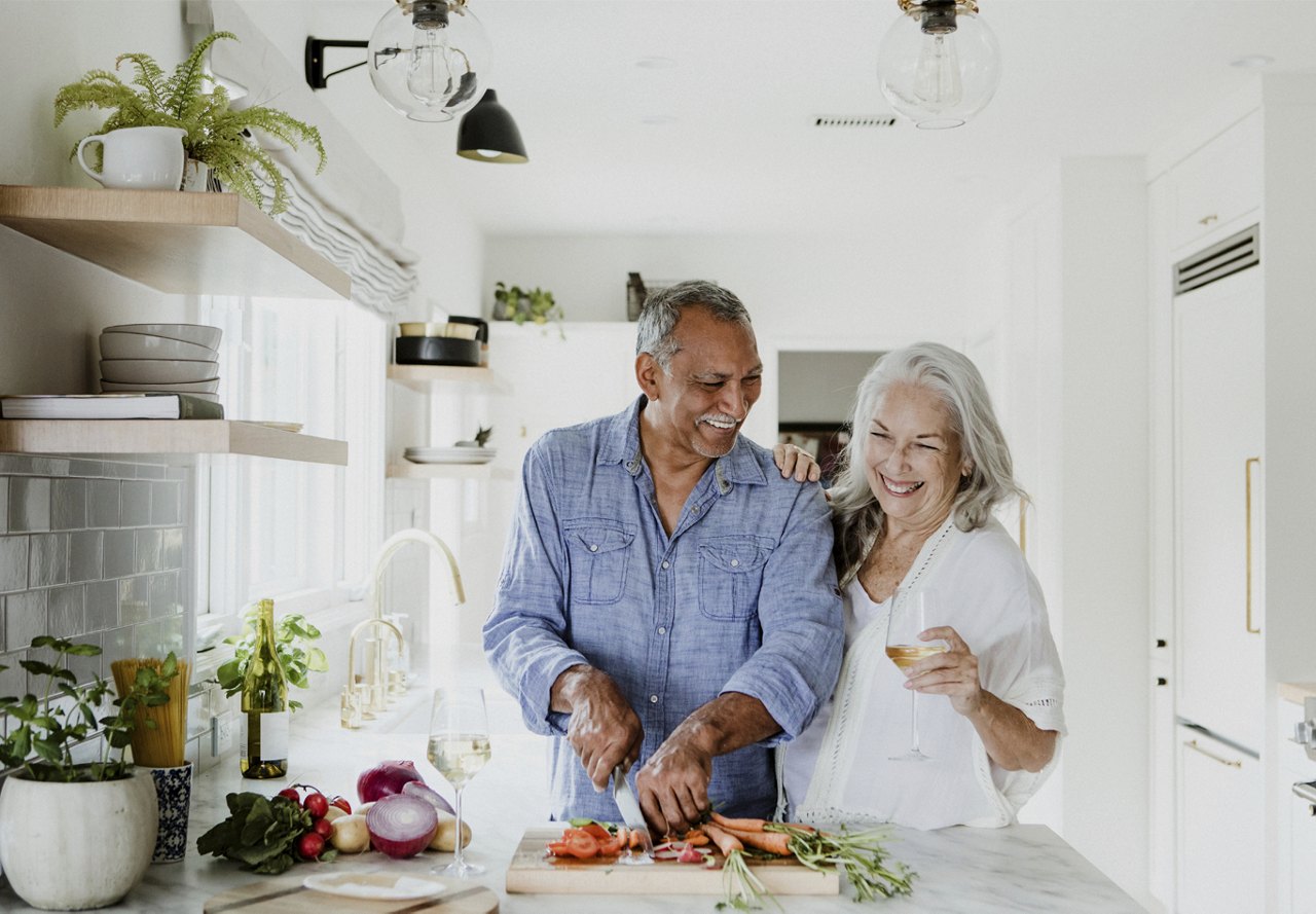 An older couple enjoying time together in their kitchen. The husband is cutting vegetables while the wife relaxes with a glass of wine. The kitchen features neutral design elements with a touch of greenery, creating a warm, inviting space.