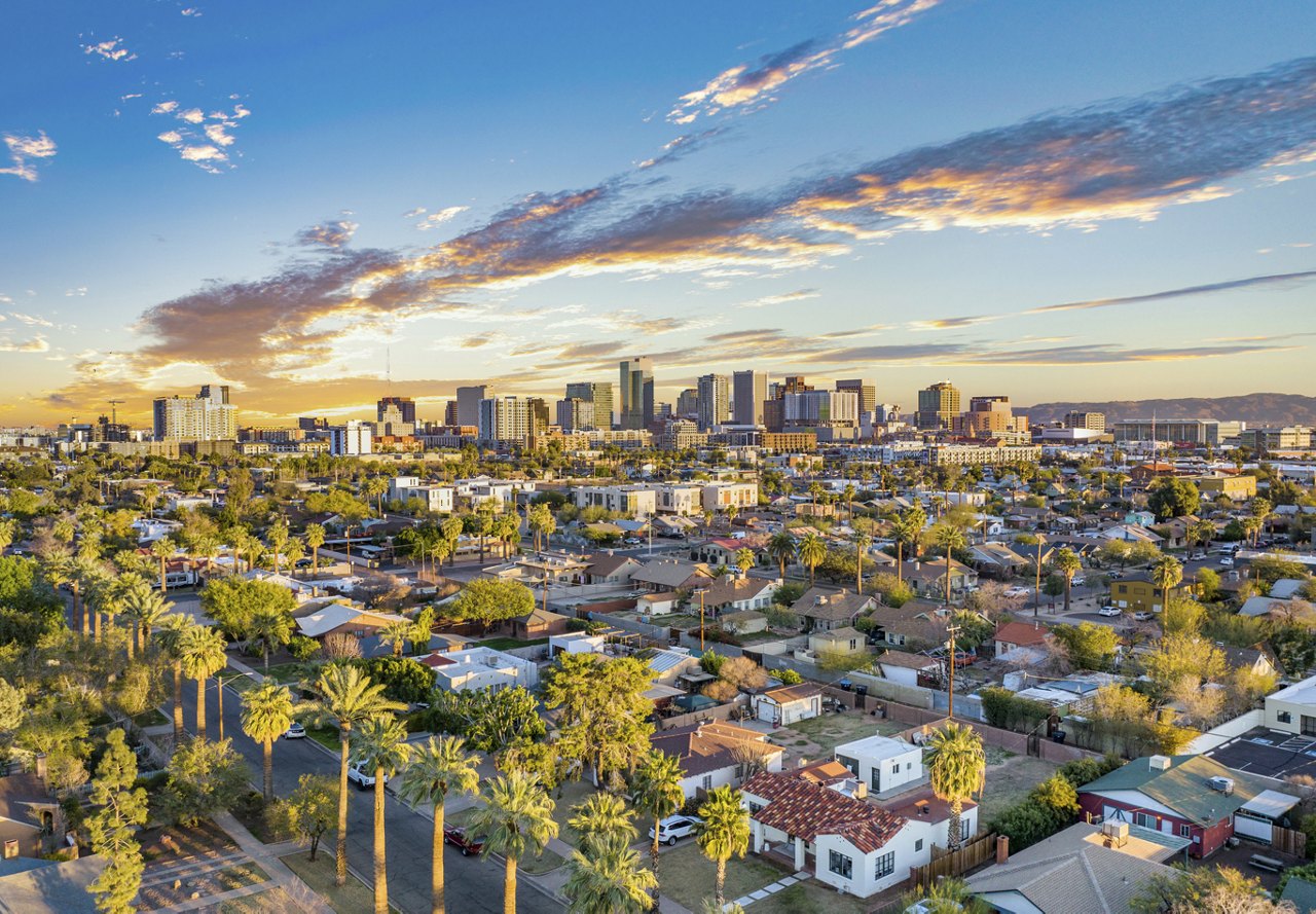 A view of Phoenix, Arizona, with a beautiful sunset casting vibrant colors across the sky. Palm trees are intertwined with the buildings below, creating a striking contrast against the clouds.