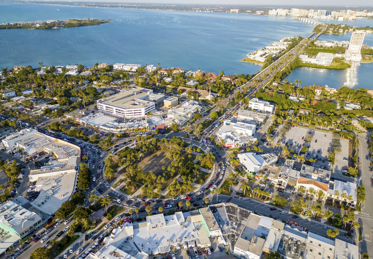 A view of Sarasota, Florida, featuring a large roundabout, water, and lush greenery. The scene highlights the beauty of the area, with buildings in the foreground and distant structures adding to the cityscape.