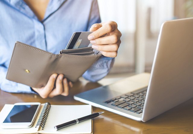 A woman pulling a credit card out of her wallet, with her computer, phone, pen, and notebook on the table in front of her as she makes an online payment.