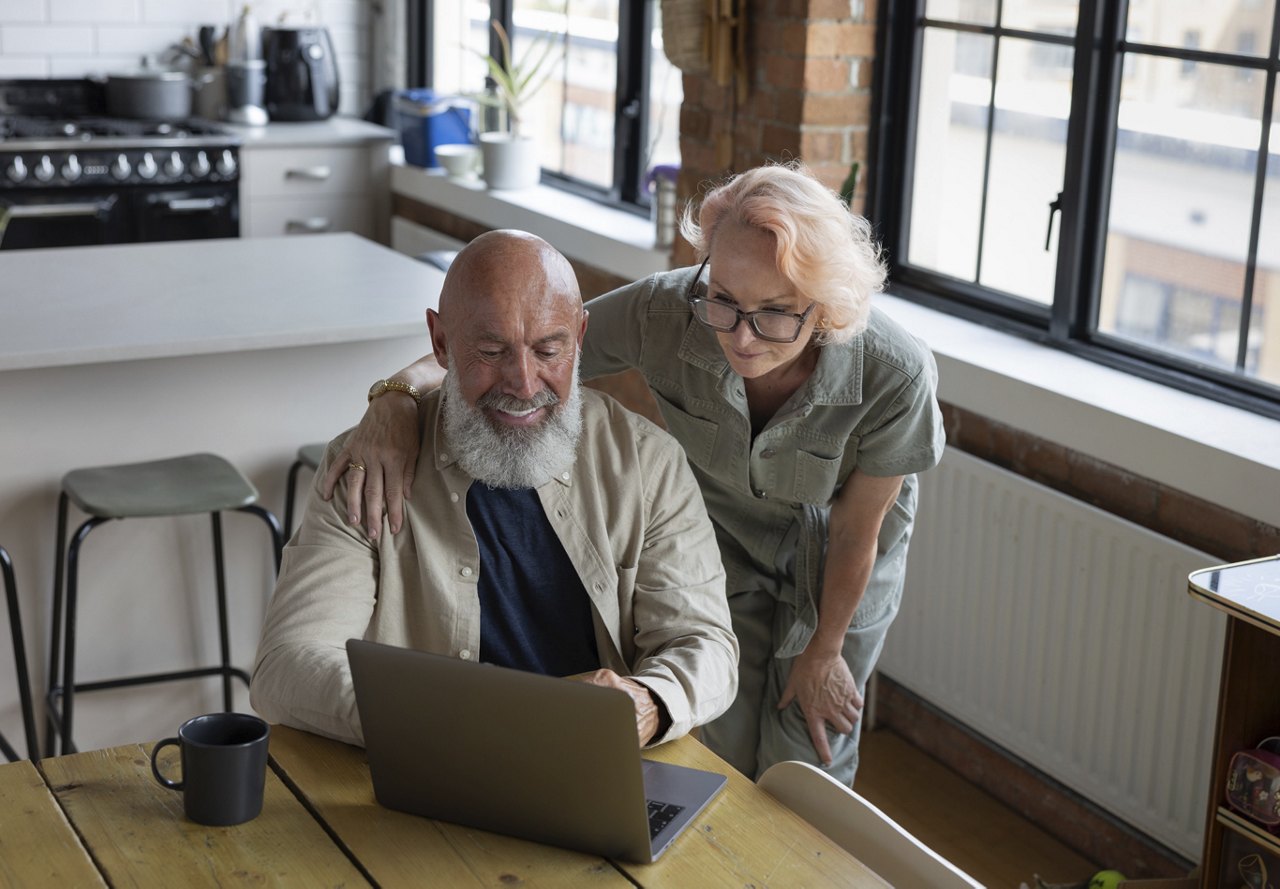 An older couple sitting together, smiling as they look at a computer while paying their bills. A coffee mug sits on the table, and the warm light from the window illuminates the kitchen area and the cozy apartment in the background.