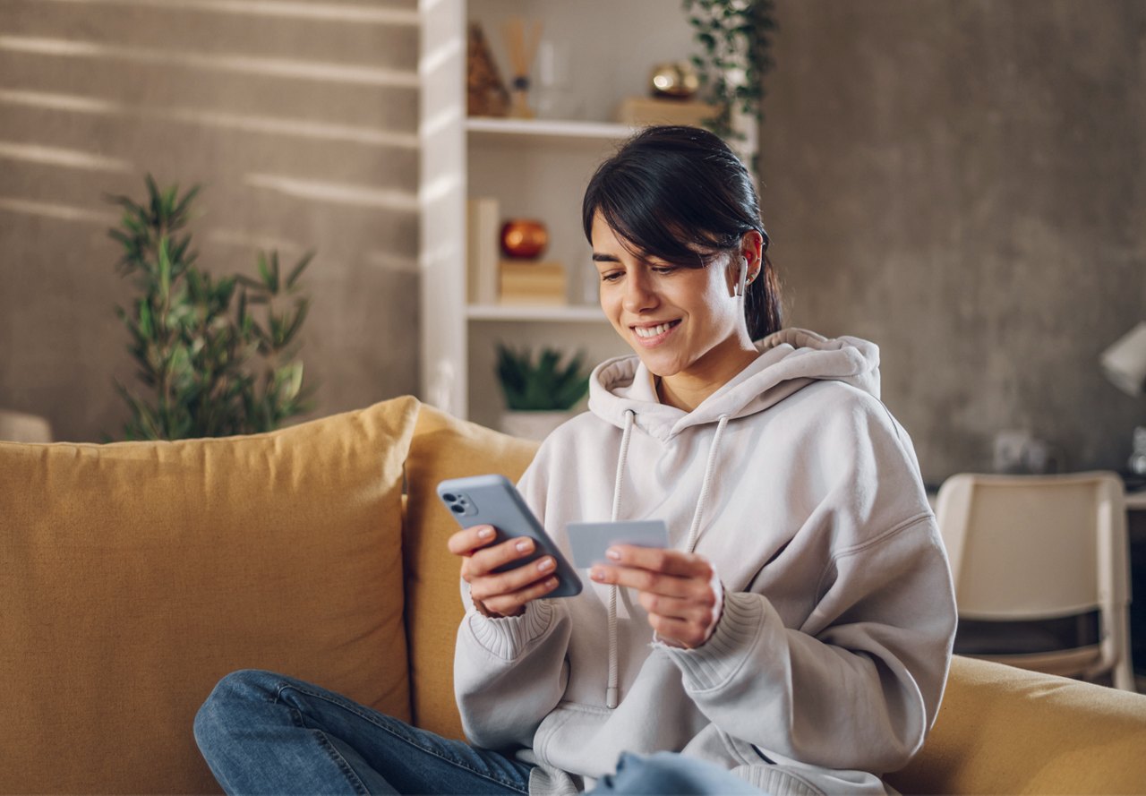 A woman sitting on the couch with her credit card in hand, preparing to make a payment on her phone. The background features earthy, inviting apartment decor, adding warmth to the space.