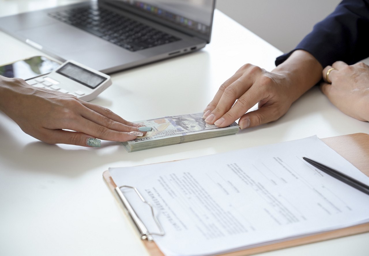 A person handing over a stack of cash to an apartment employee to pay their security deposit. Nearby, you can see the signed paperwork, a calculator, and a computer on the desk.