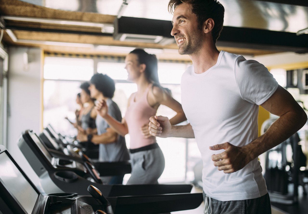 A man smiling while running on a treadmill in his apartment, with large windows letting in natural light. In the background, several people are running, blurred out, creating a sense of movement.