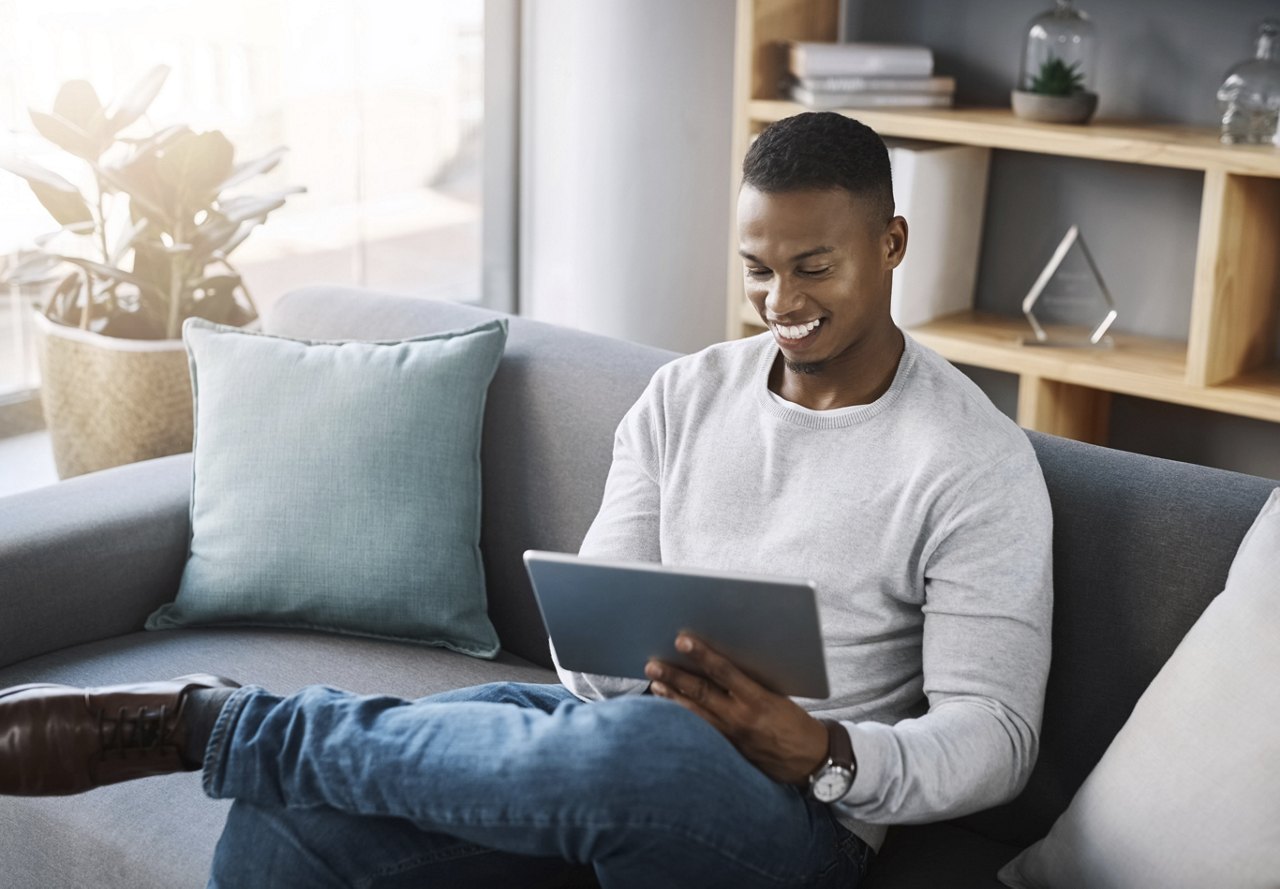 A man sitting on a couch in his home, smiling as he uses a tablet. Natural light from the window illuminates the space, creating a warm and inviting atmosphere.