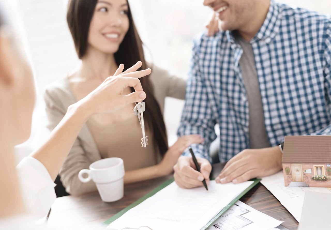 A couple looking at each other as they sign a lease, with a rental employee handing them a set of keys to their new home.