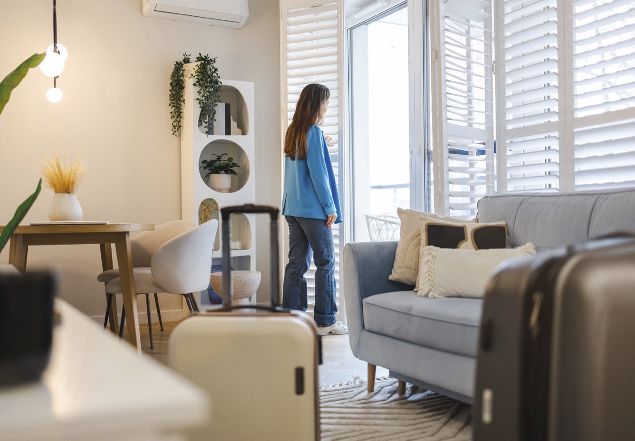 A woman standing at the door of her apartment, looking out with a suitcase in the foreground, suggesting she’s about to travel. The room is filled with natural light, showcasing the home’s decor.