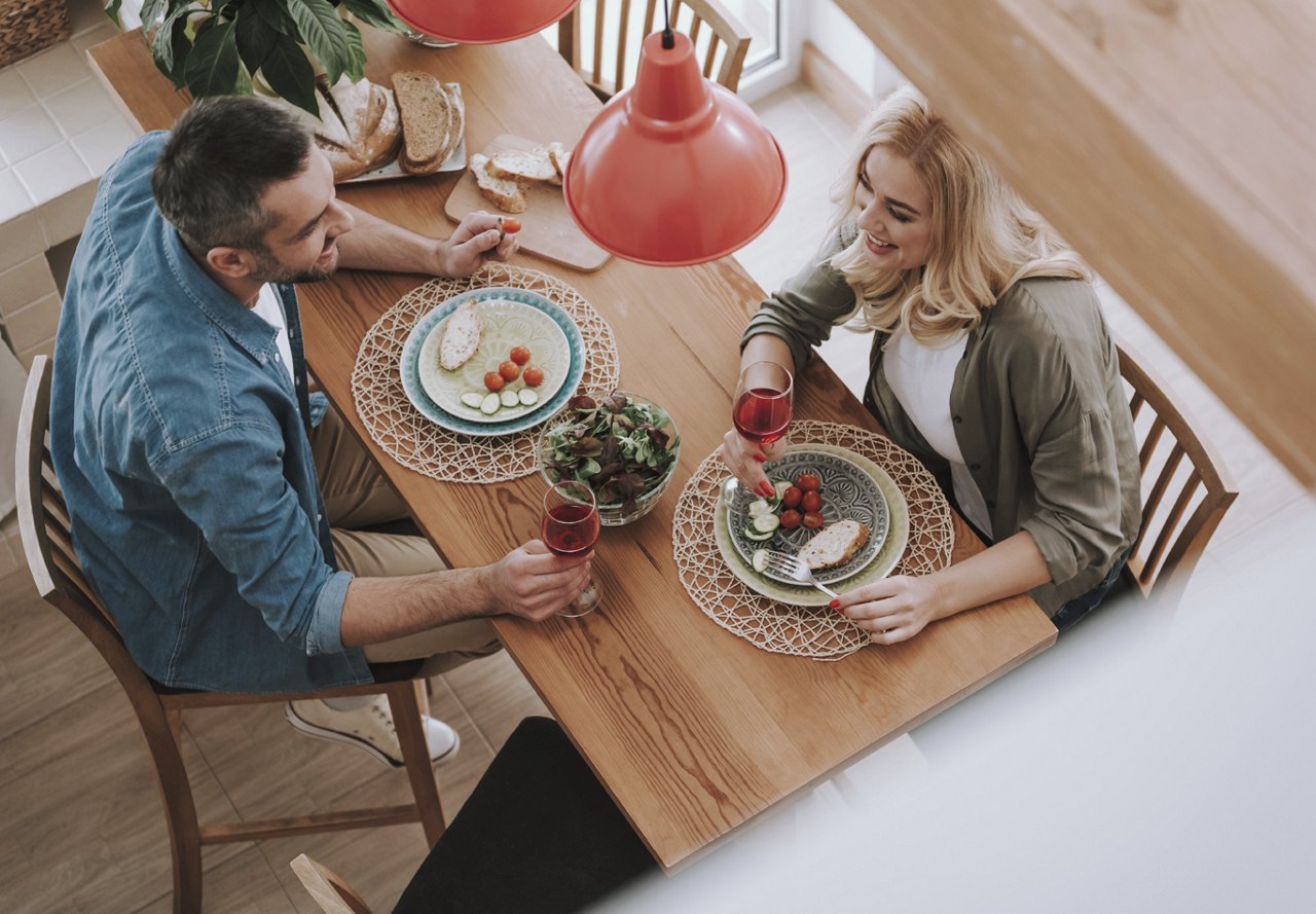 A couple sitting at a dining table, enjoying a meal and conversation, smiling and having a good time. The image is taken from above, capturing their relaxed and joyful moment.