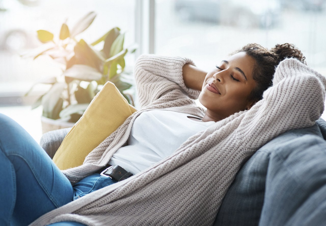 A woman relaxing on the couch with her hands in her hair, a large window behind her filling the space with natural light. An indoor plant adds a touch of greenery to the room.