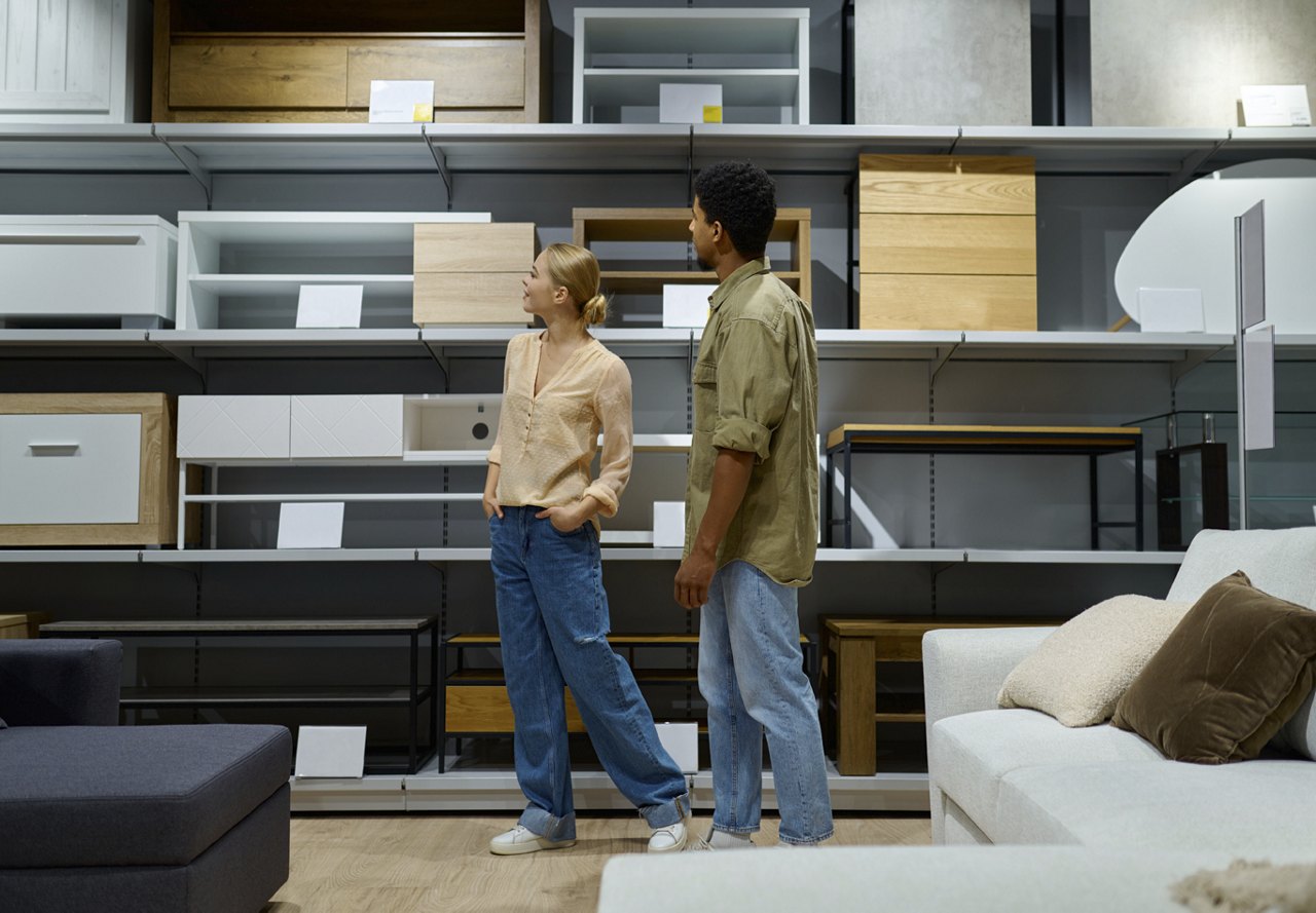 A couple strolling through a well-lit furniture store, thoughtfully browsing for new pieces to add to their home. Behind them, shelves are neatly stocked with a variety of coffee tables.