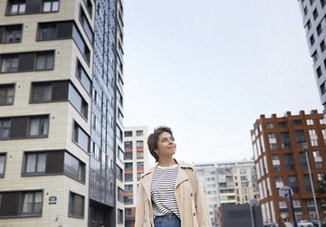 A woman walking through her neighborhood, looking up at the towering skyscrapers above her. The clear sky adds to the vibrant, urban atmosphere of the scene.