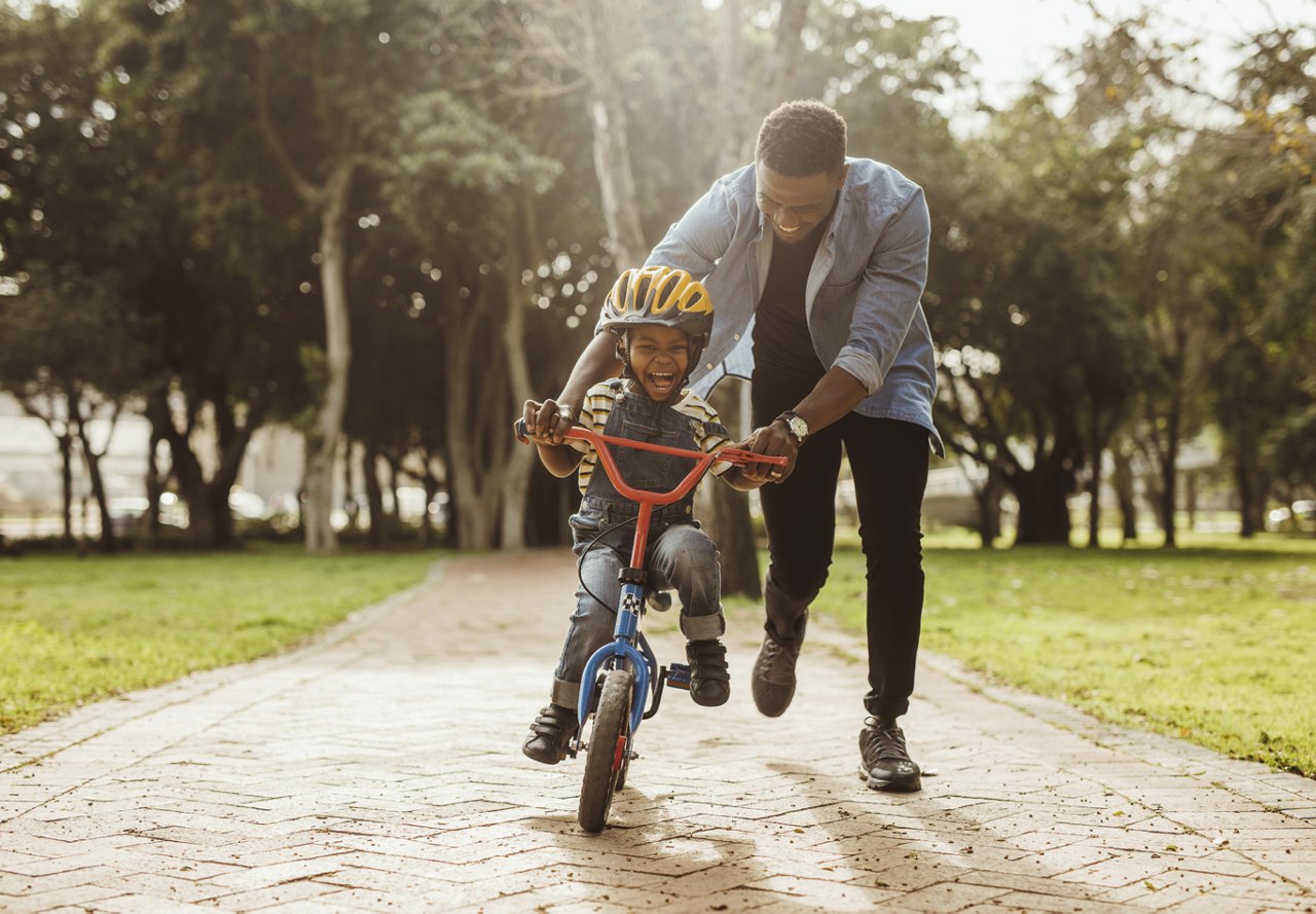 A man and his son at a park, with the son learning to ride a bike. Both are smiling, enjoying the beautiful day together in the outdoors.