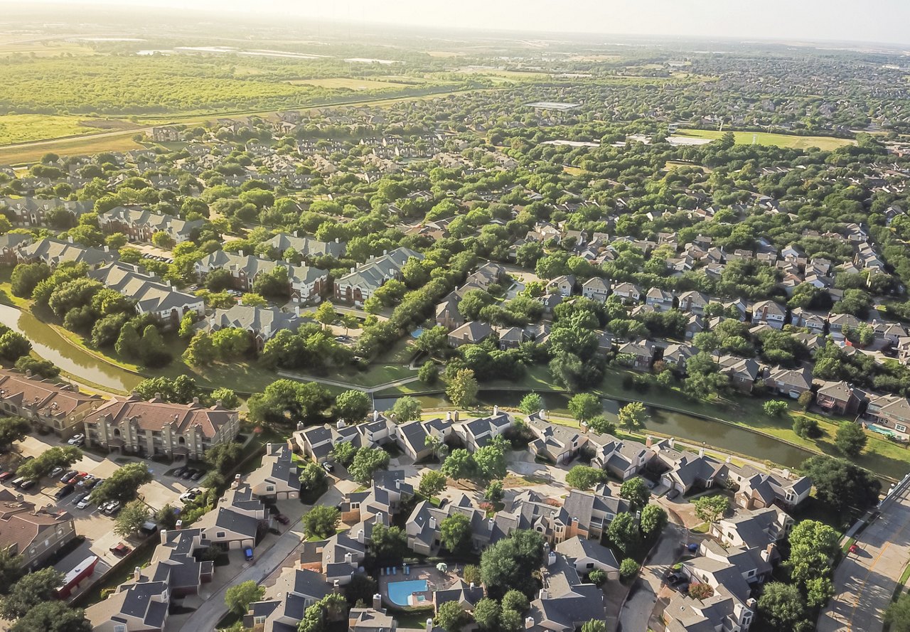 An aerial view of a suburban neighborhood, featuring bright green lawns and numerous houses, with the sun shining down, highlighting the peaceful, open atmosphere.