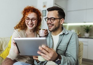 A couple sitting together on a couch in their home, smiling as they look at a tablet with coffee in hand while searching for a new apartment.