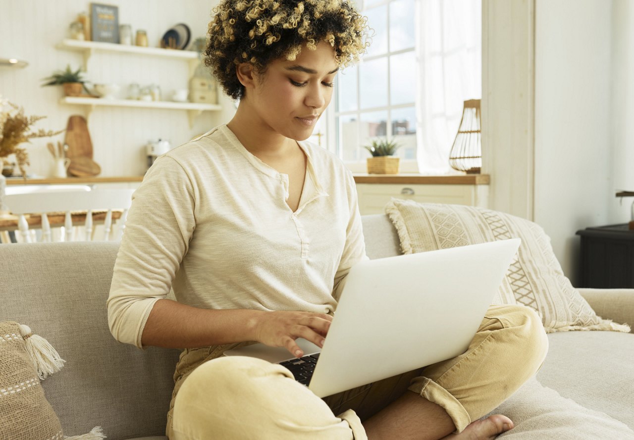 A woman sitting on the couch in her apartment, looking at the computer on her lap as she researches apartment options, with a view of her kitchen in the background.