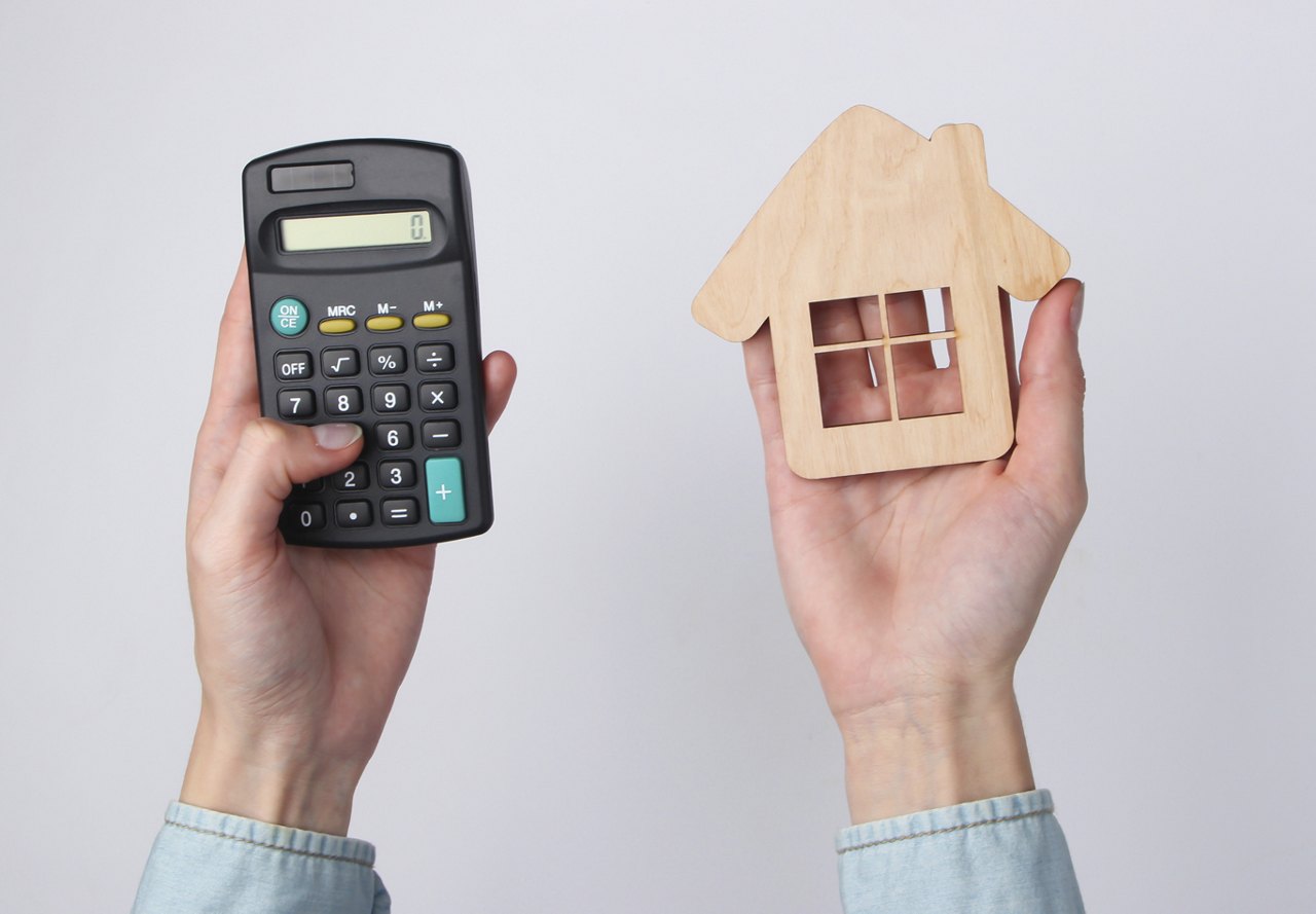 Two hands, one holding a calculator and the other holding a small wooden house, representing the process of calculating home costs.