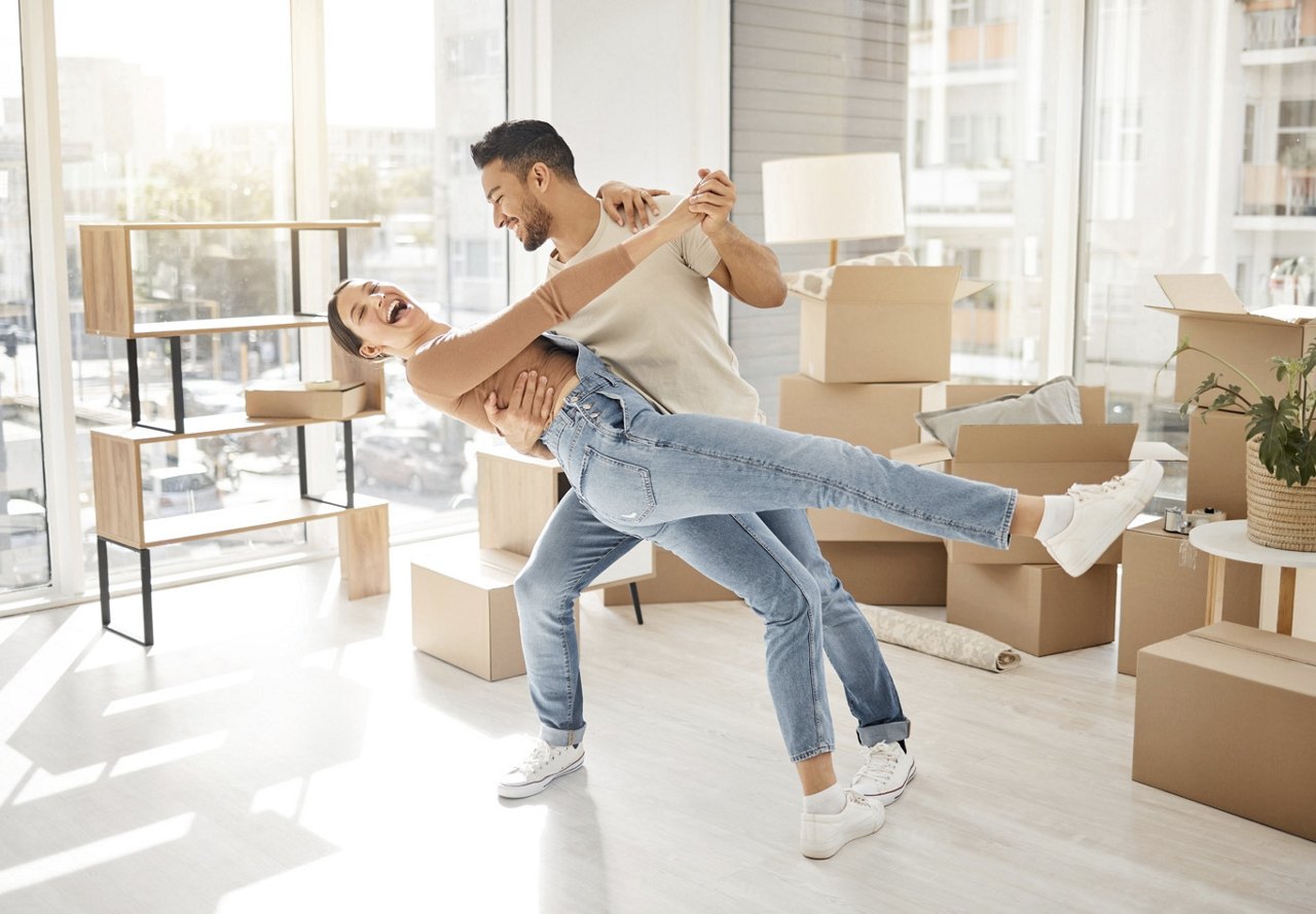 Happy couple dancing in their new home among moving boxes, with the man playfully dipping the woman as sunlight streams through a large window.