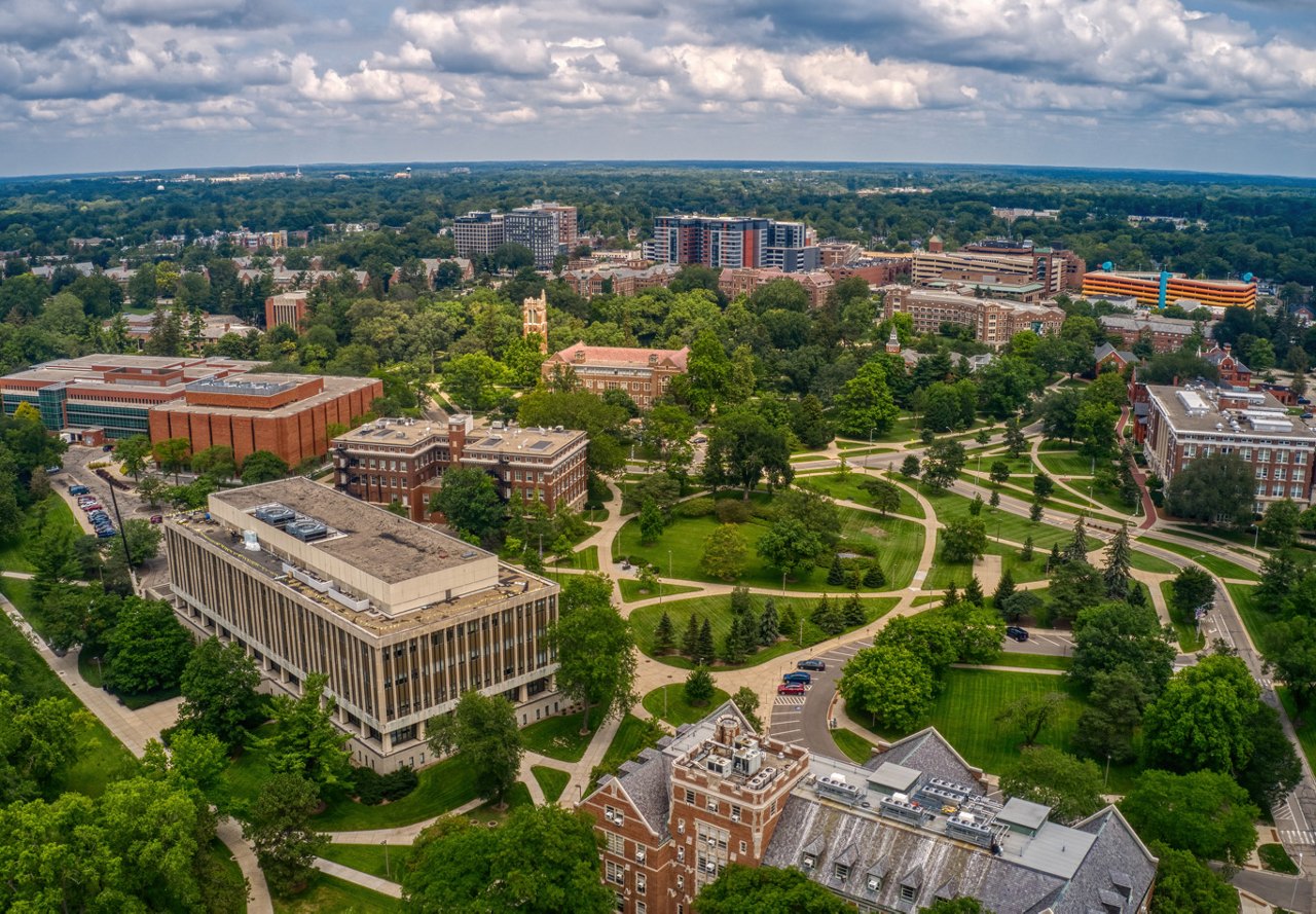 An aerial view of a picturesque college campus featuring academic buildings, winding walking paths, and lush greenery, all framed by a vibrant sky with soft, scattered clouds.