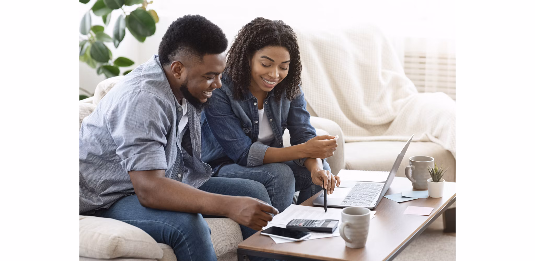 African American couple sitting on their couch, reviewing financial documents and a computer on the coffee table, with a plant in the background.
