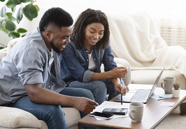 African American couple sitting on their couch, reviewing financial documents and a computer on the coffee table, with a plant in the background.