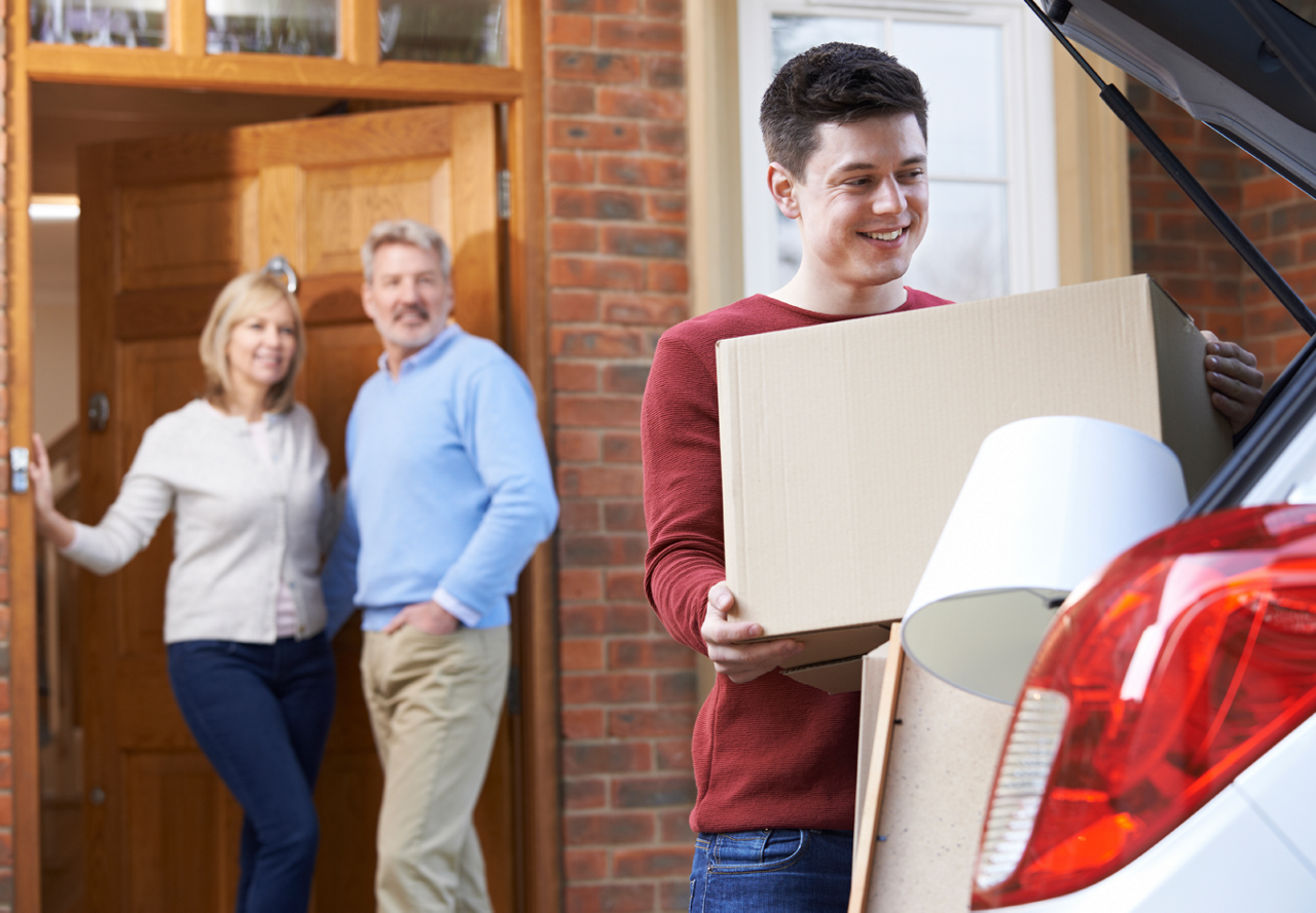 Older couple watching their son happily pack boxes into his car as he moves out for college, smiling as they see him off.