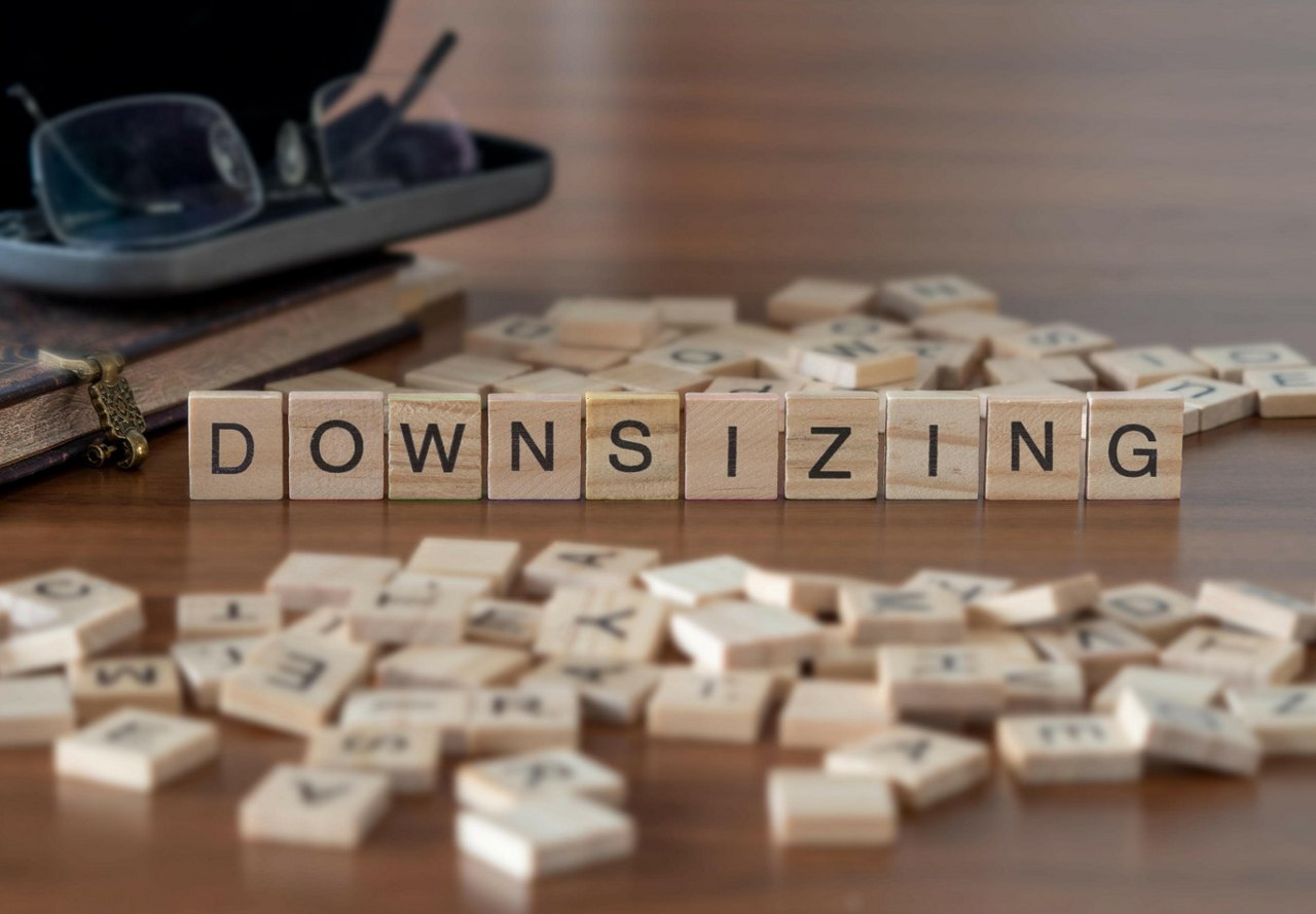 Scrabble letters on a wooden floor, with some standing upright to spell 'downsizing.' A glasses case with a pair of glasses rests in the blurred background.