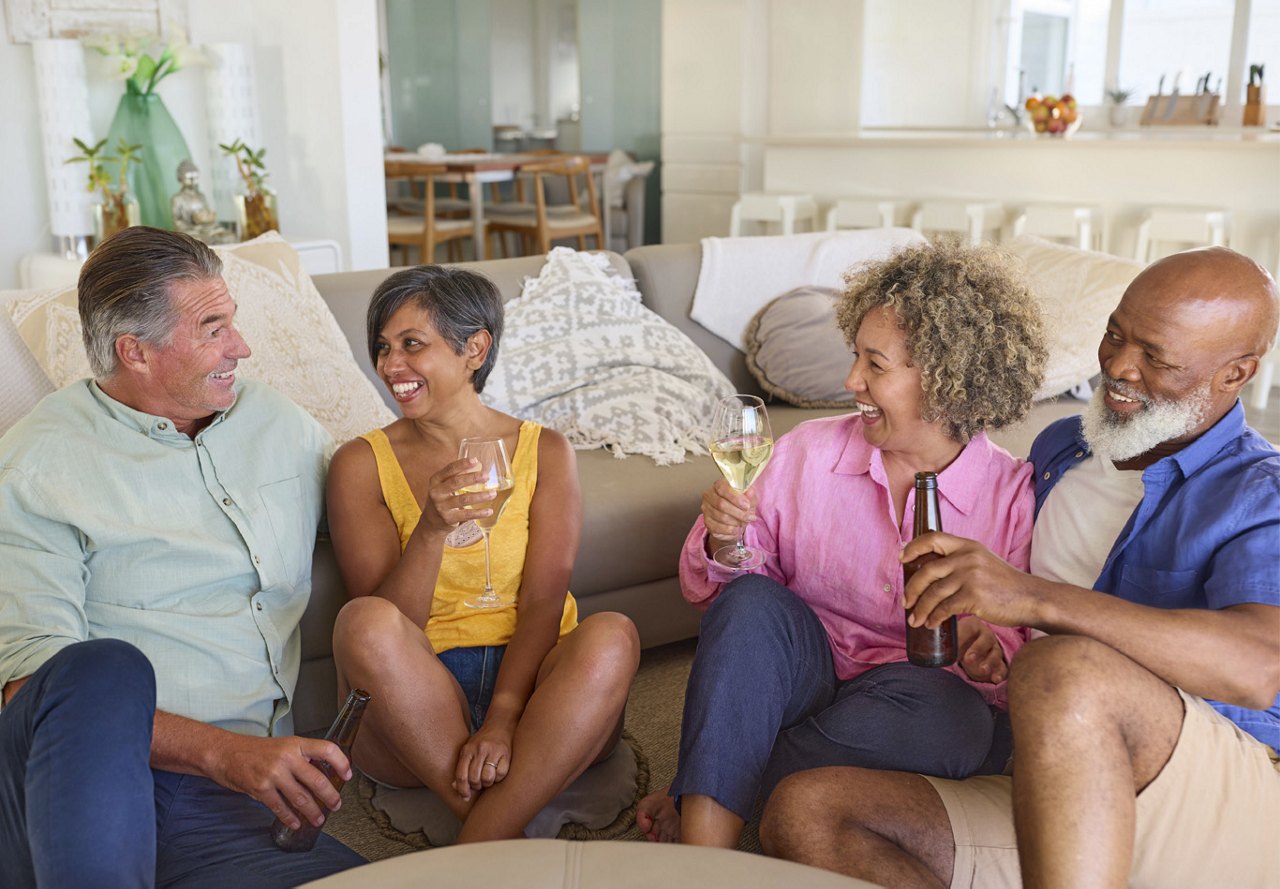 Four older adults sitting together on the floor, socializing with drinks in hand, in a warmly decorated home with vibrant colors in the background.