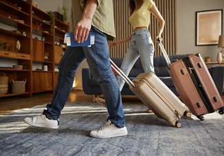 Two people rolling suitcases through their home while leaving for vacation, one holding boarding passes and passports, with a view of their stylish living space in the background.