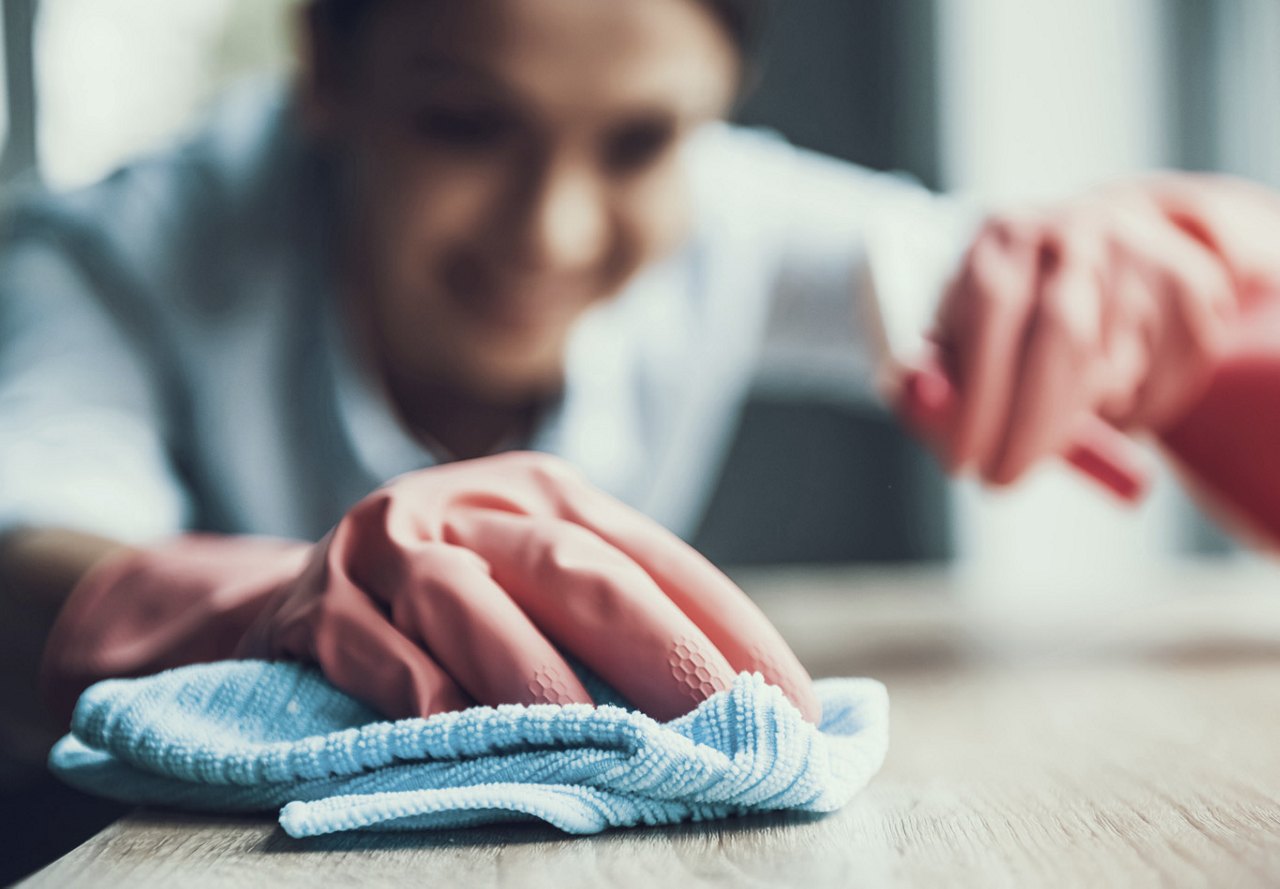 Close-up of a gloved hand wiping a surface with a rag, while a woman is blurred in the background in her apartment home, focusing on cleanliness and care.