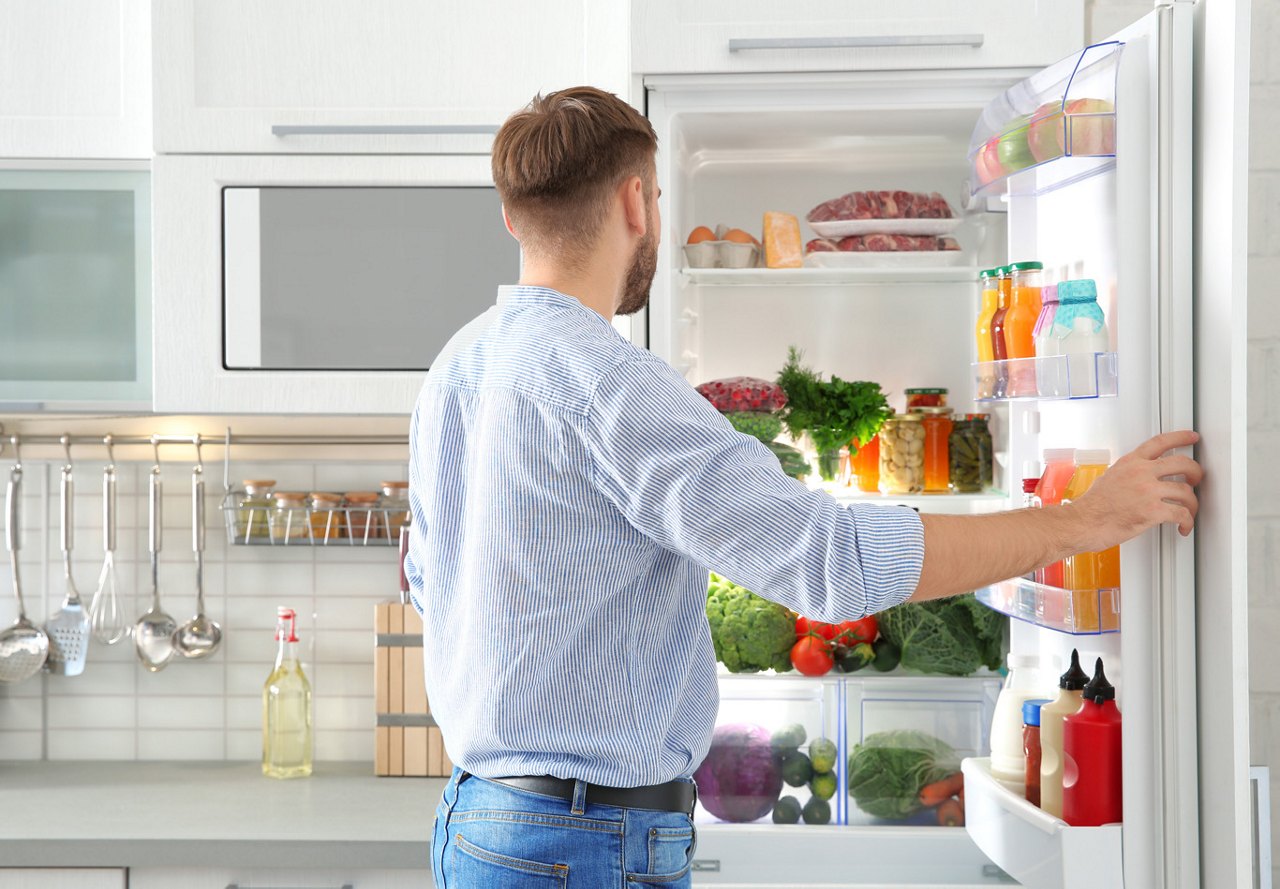 Man looking into a fridge filled with colorful fruits and vegetables, with a view of his kitchen on the left, showcasing a well-organized and vibrant home.