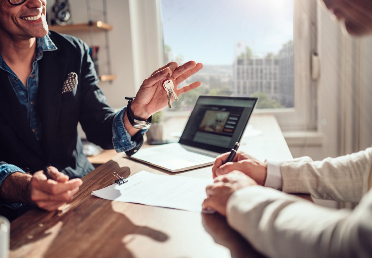 People sitting at a desk, speaking with a rental agent who is holding out keys to their new home with a smile, while a city view is visible through the window in the background.