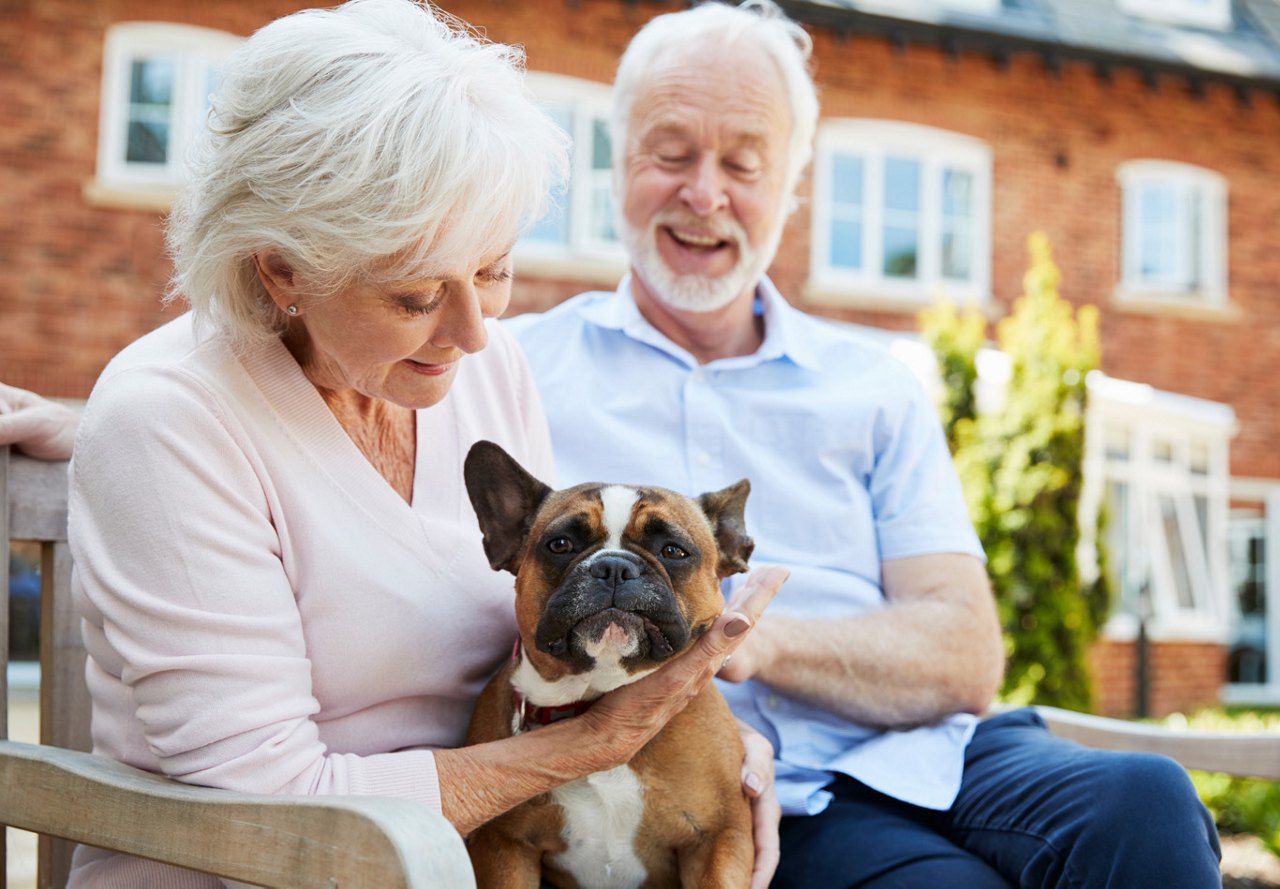 An older couple sitting together on a bench outside their senior living facility, smiling warmly while their dog sits beside them, surrounded by a peaceful outdoor setting.