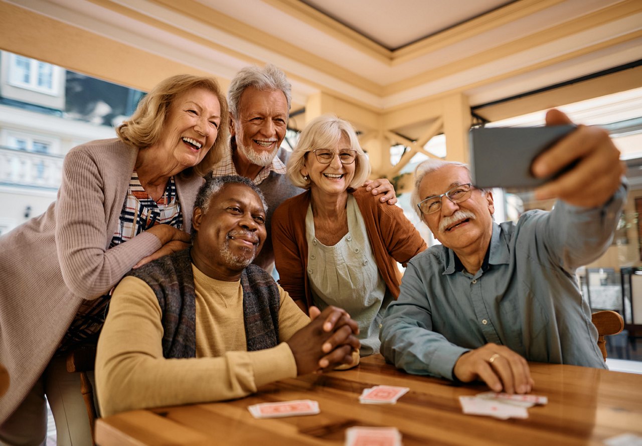 A diverse group of five older individuals sitting together at a table, smiling and taking a selfie while playing cards and enjoying each other's company.