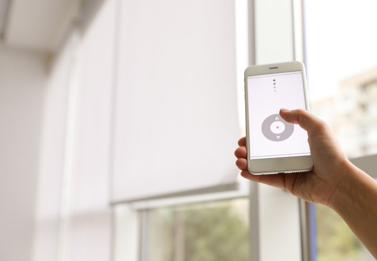 Person holding a white remote while using a smartphone app to control automatic blinds, with a blurred view of the outdoors visible through the windows in the background.