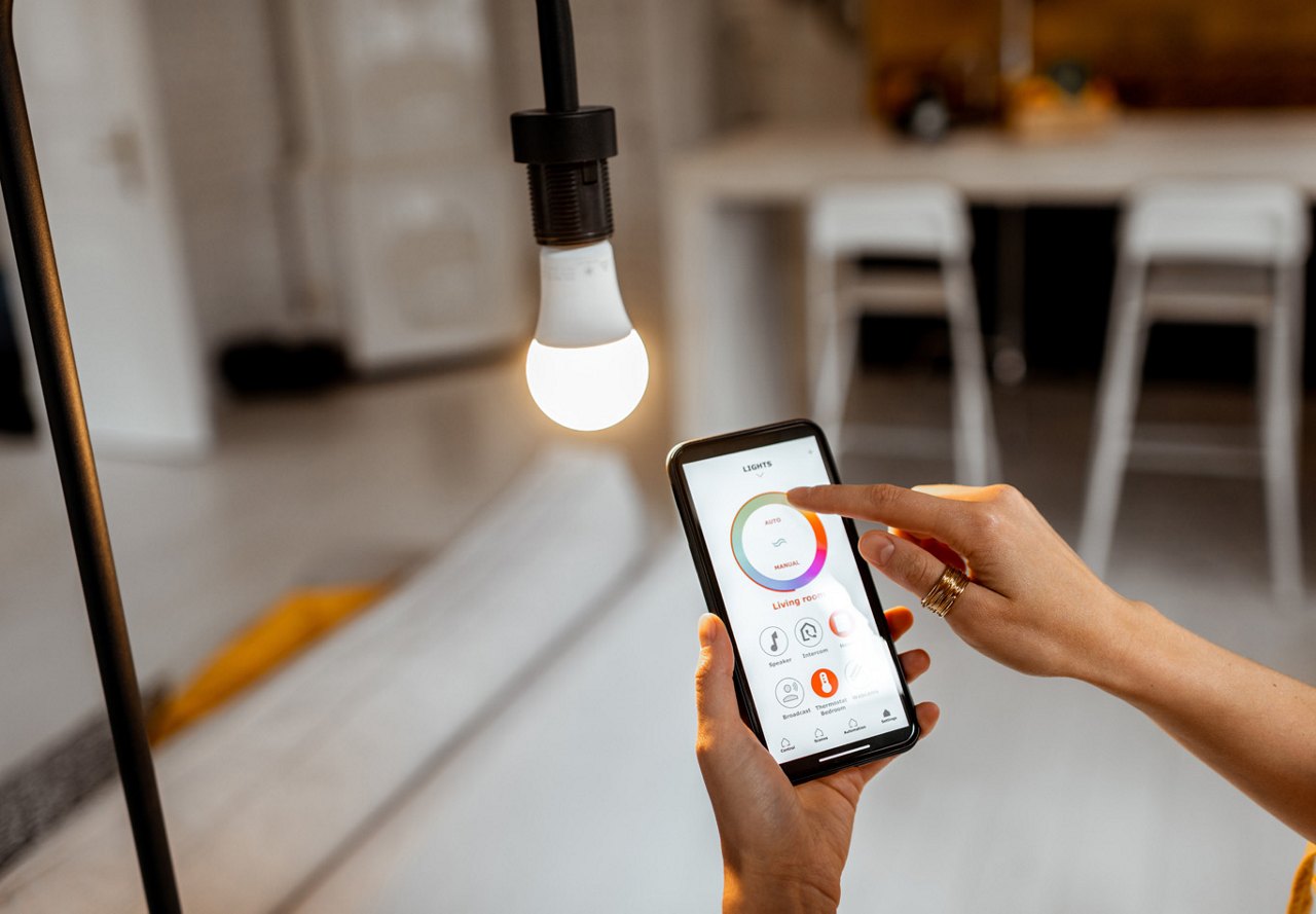 erson using a smartphone to adjust a brightly lit lightbulb in their modern apartment, with a softly blurred background showcasing cozy home decor.