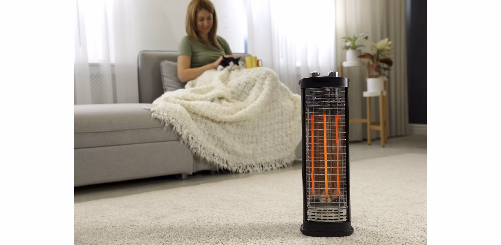 A woman sitting on her couch wrapped in a blanket, cuddling her pet cat, while a space heater warms the living area during the winter months. The cozy atmosphere is enhanced by house plants in the background.