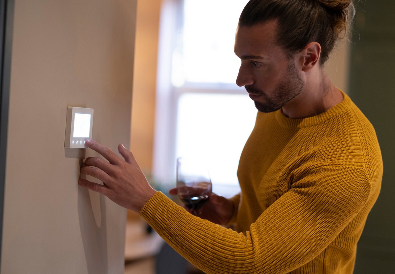 A man with a ponytail adjusting the thermostat in his home to make it warmer during the cold months, while sipping on wine and relaxing in a cozy yellow sweater.