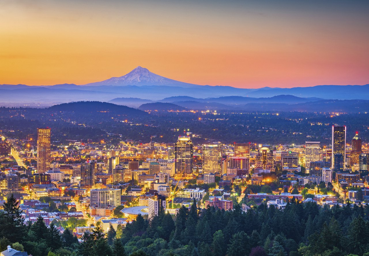 A stunning view of Portland, Oregon, with a colorful sunset, city lights below, and distant mountains adding to the vibrant cityscape.