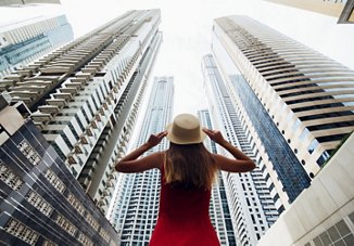 A woman in New York City holding onto her hat as she looks up in awe at the towering skyscrapers above her.