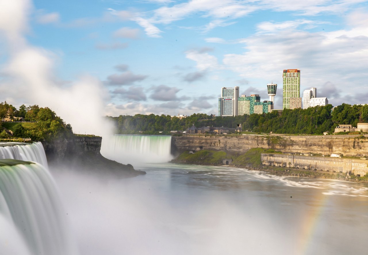 A stunning view of Niagara Falls in Buffalo, New York, with a rainbow in the foreground and city buildings visible in the distance.