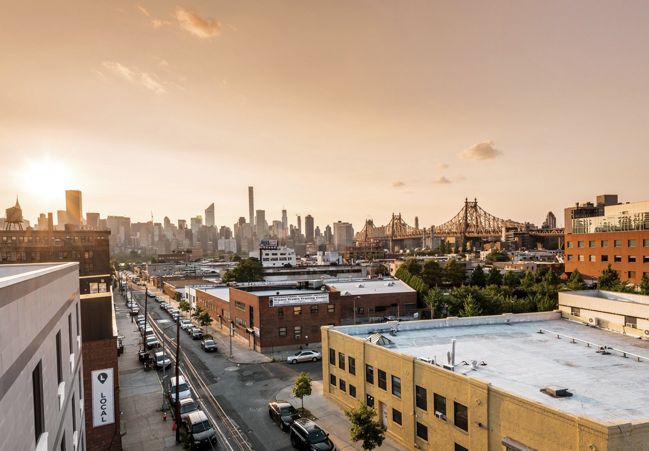 A scenic view of a Queens neighborhood in New York, featuring the Brooklyn Bridge and the Manhattan skyline in the distance.