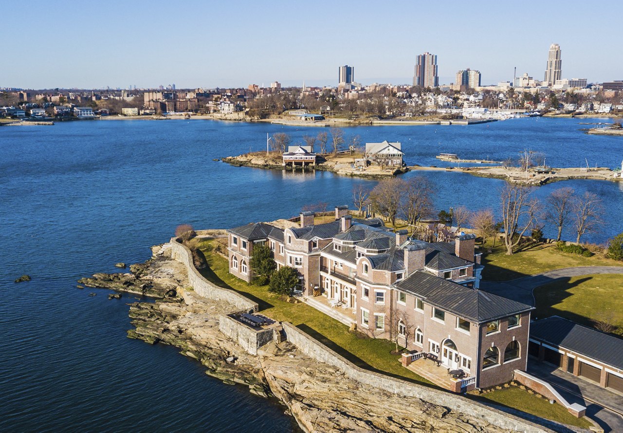 A scenic view of the waterfront in Westchester, New York, featuring buildings along the shore, lush greenery, and a clear blue sky, with a city skyline visible in the background.