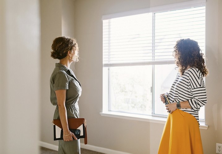 A woman tours an empty apartment, looking out a large window while the real estate agent observes her reaction.