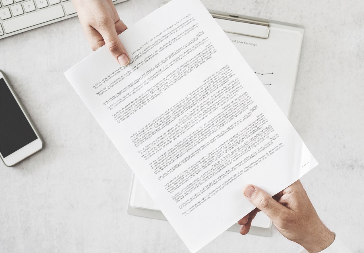 A person hands a document with text to another individual across a desk, which also has a clipboard, cell phone, and keyboard.