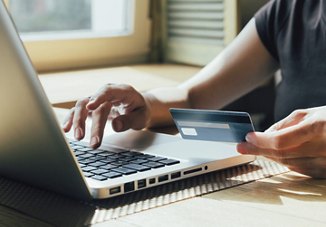 A person holding out their credit card and looking at it while typing on a computer to digitally pay their rent, with light streaming through the window behind them and a blurred background.